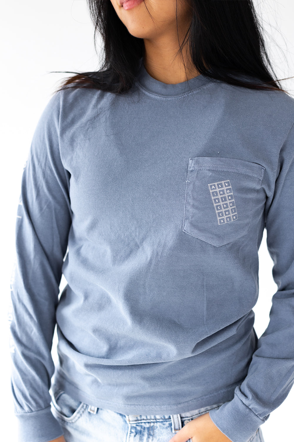 Person wearing a blue-grey long-sleeve shirt with a chocolate bar on the pocket and Askinosie Chocolate text on sleeve against a white background