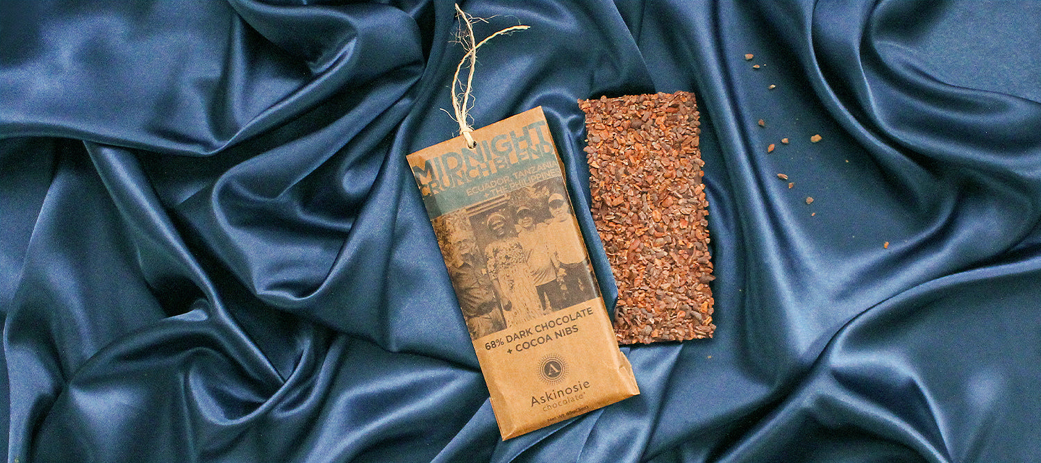 Kraft brown packaging next to a nib-coated chocolate bar laid on a silk blue background