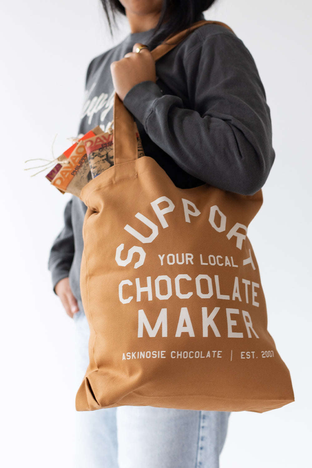 Person holding a brown tote bag with 'Support Your Local Chocolate Maker' text, wearing a gray sweatshirt.