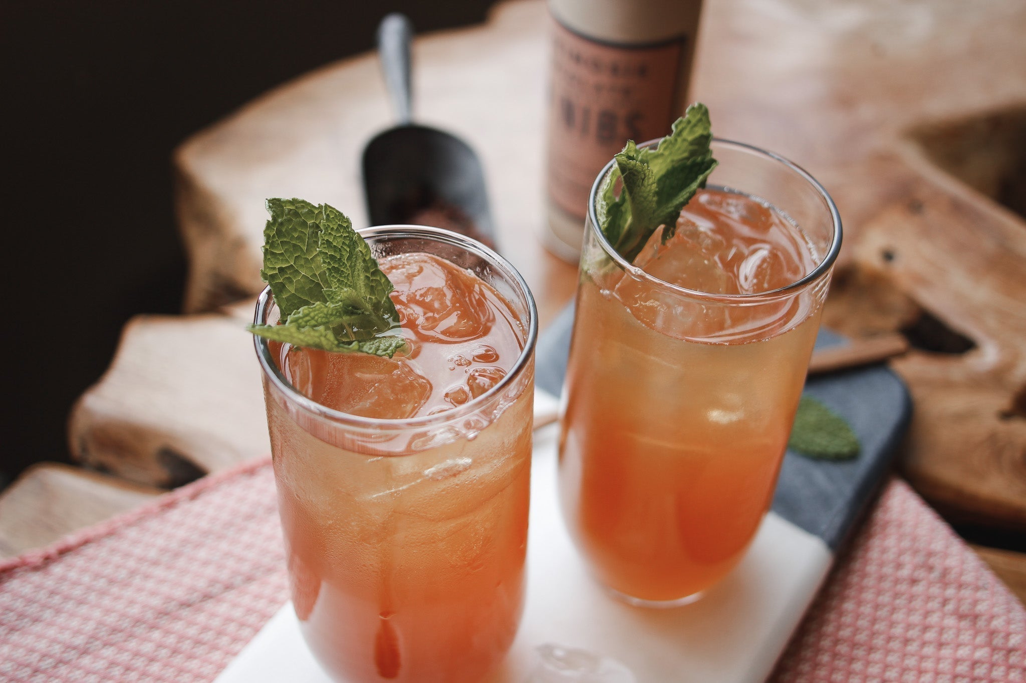 Two cups of Iced Nib Tea sit on a marble serving tray with a cotton placemat on top of a wooden table.