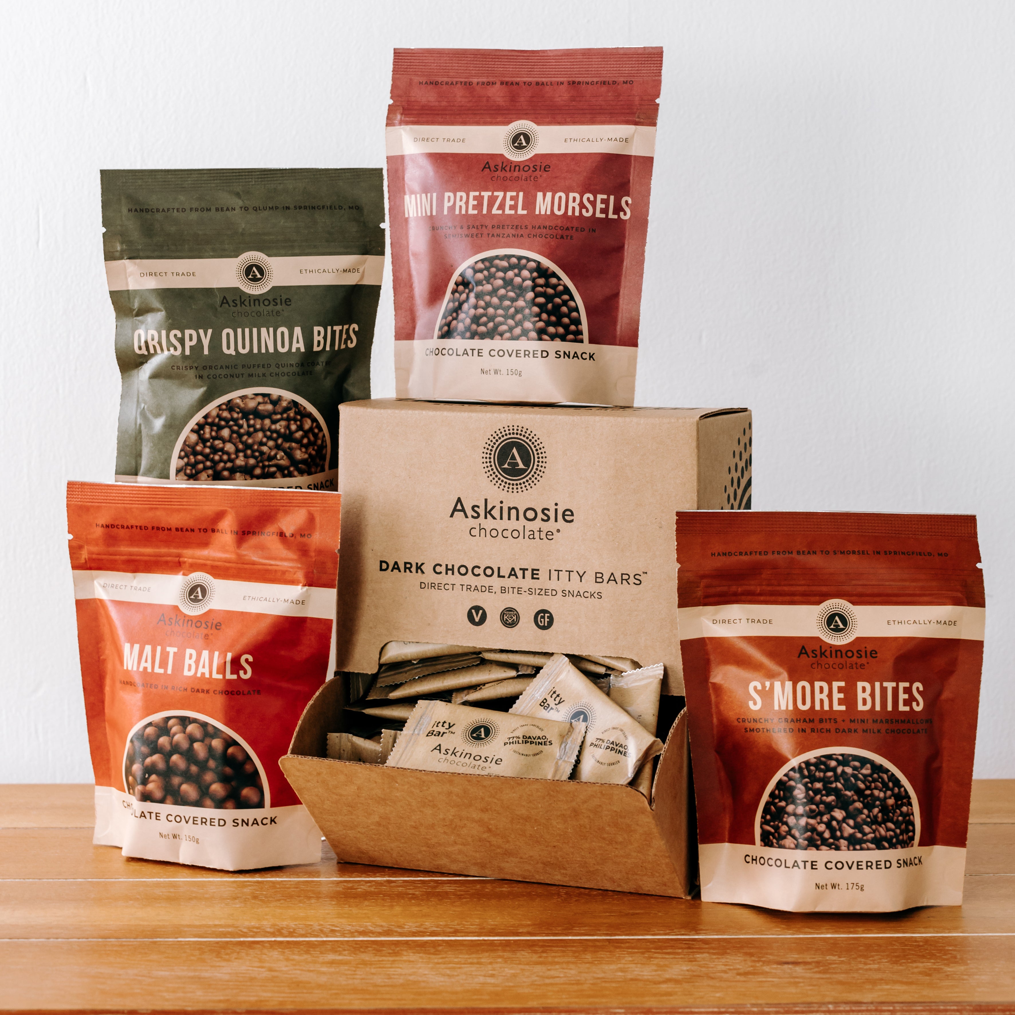 Jewel-toned kraft pouches against a white background with a kraft display box full of bite-sized chocolate bars in the center.