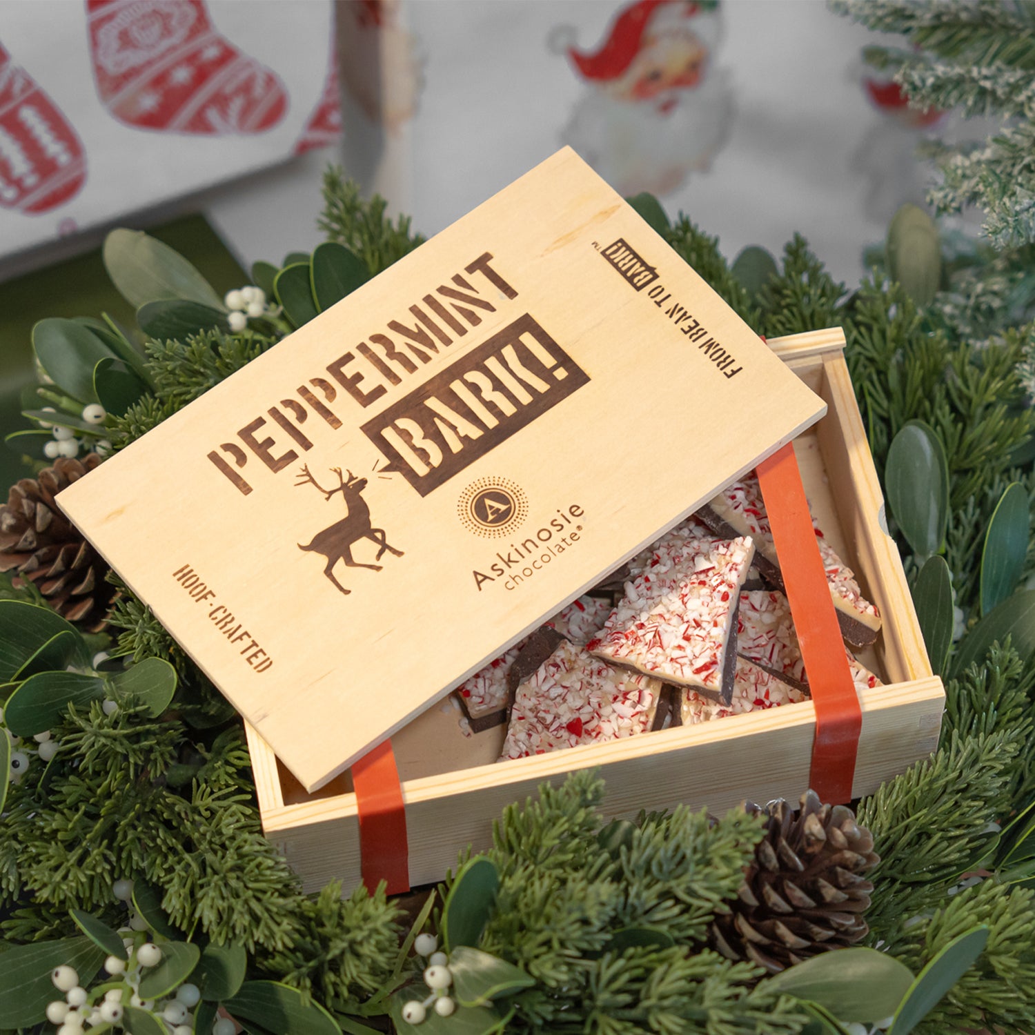 A pine box of Peppermint Bark slightly open to show the chocolate inside, against a background of greenery