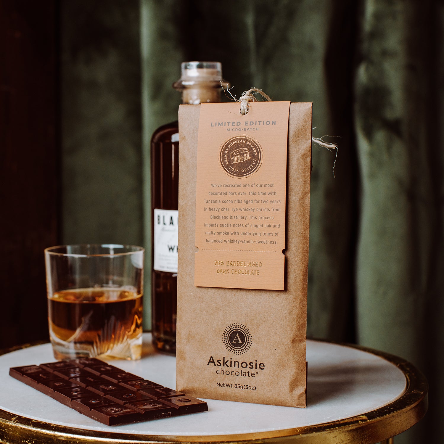 Askinosie Chocolate Limited Edition Barrel-Aged Dark Chocolate Bar, bottle of whiskey, and glass on a marble and gold table with a blurred background