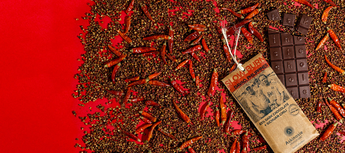 A dark brown chocolate bar and kraft packaging laid on top of dried chili peppers and peppercorns on a red background