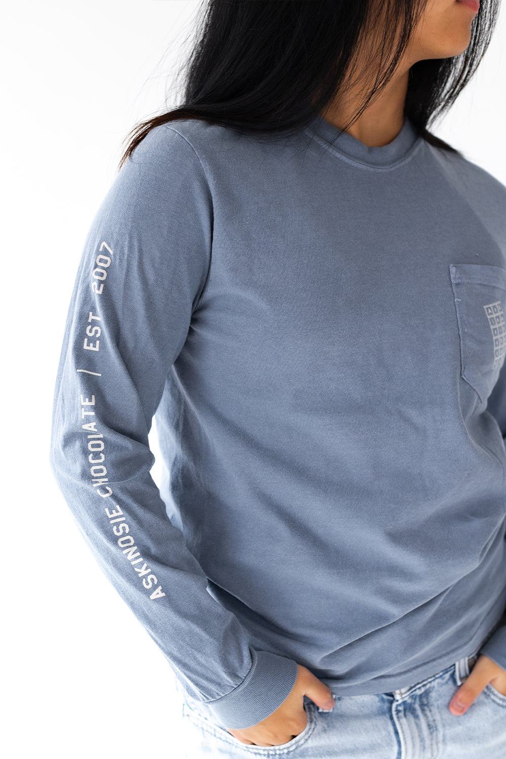 Person wearing a blue-grey long-sleeve shirt with a chocolate bar on the pocket and Askinosie Chocolate text on sleeve against a white background, facing away from the camera