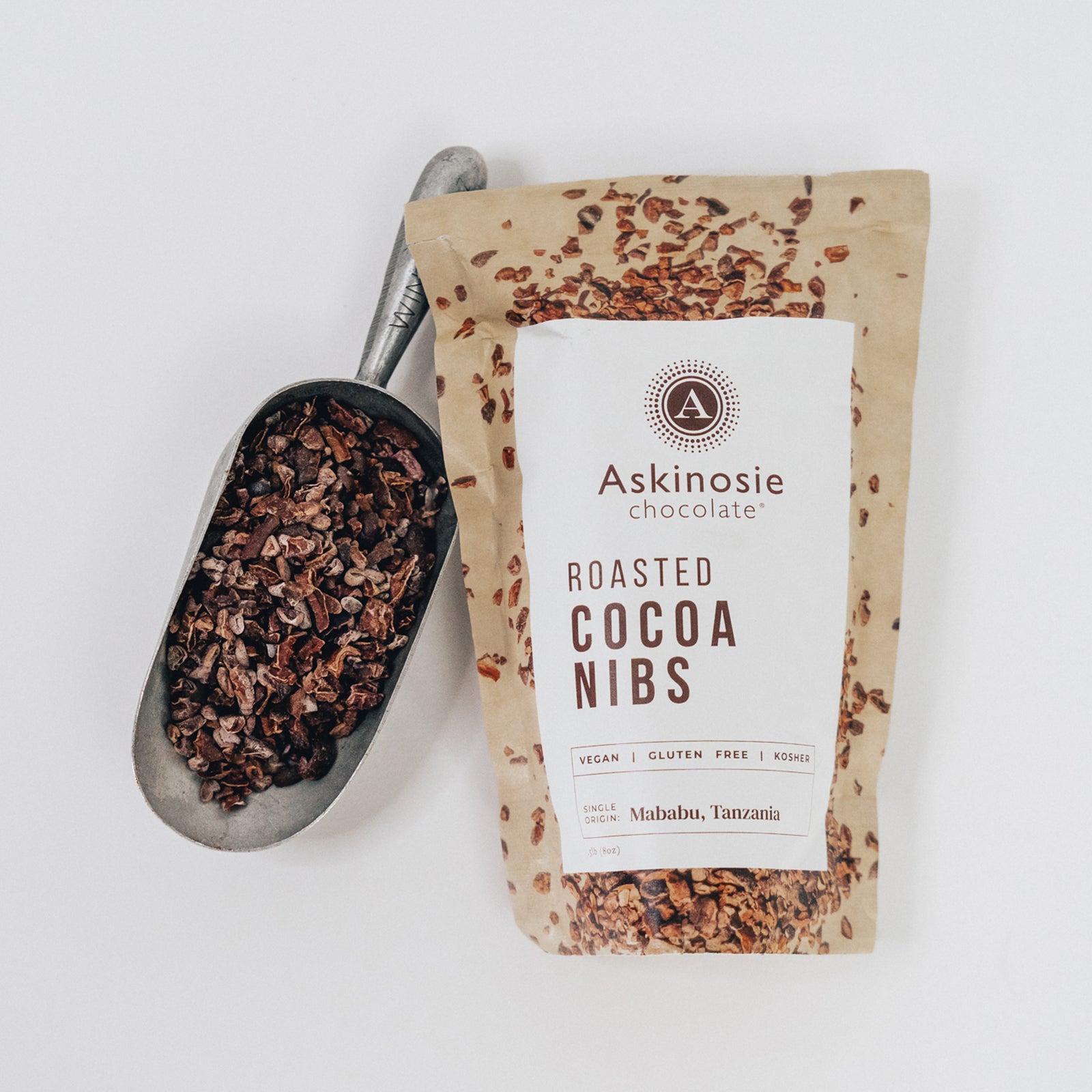 Kraft pouch of Askinosie Chocolate roasted cocoa nibs with a scoop of nibs on a white background