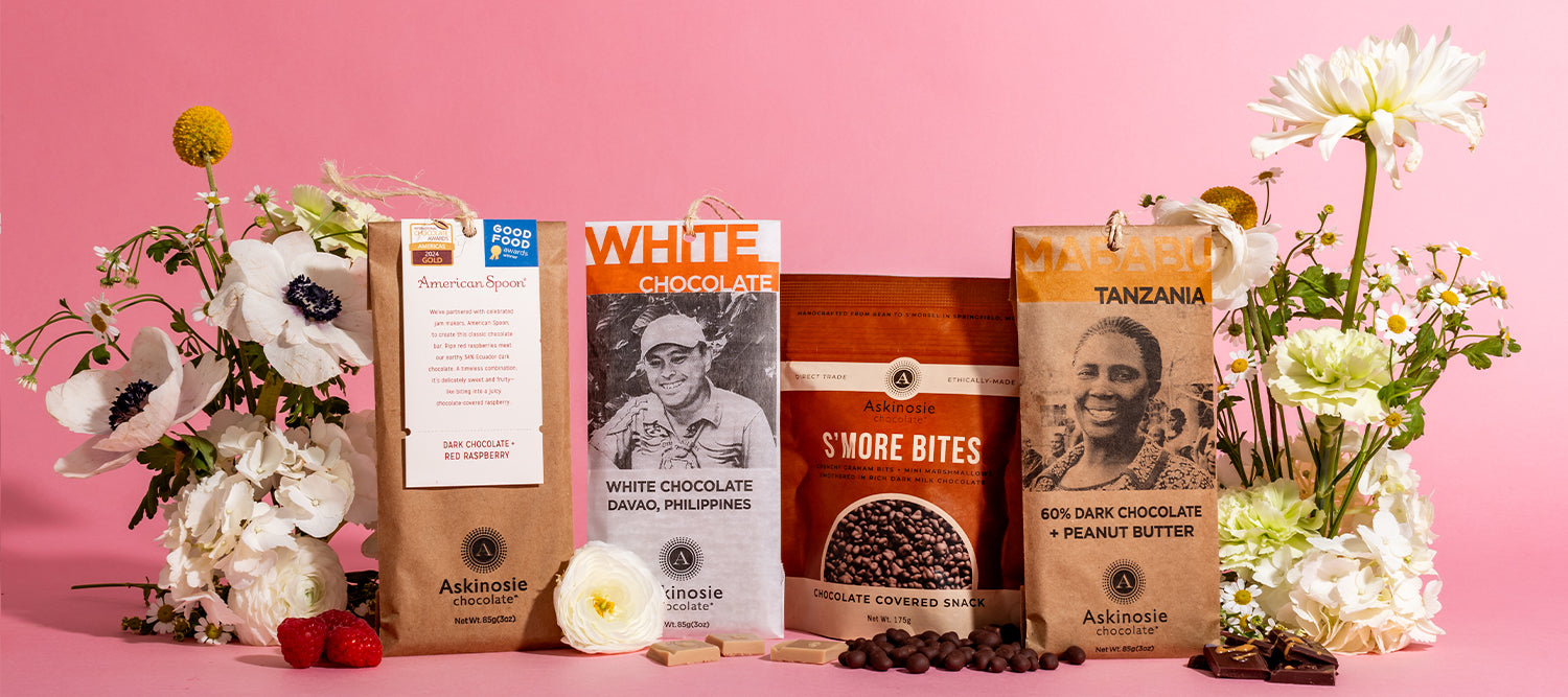 A group photo featuring several Askinosie Chocolate products arranged between two groups of flowers in front of a pink background