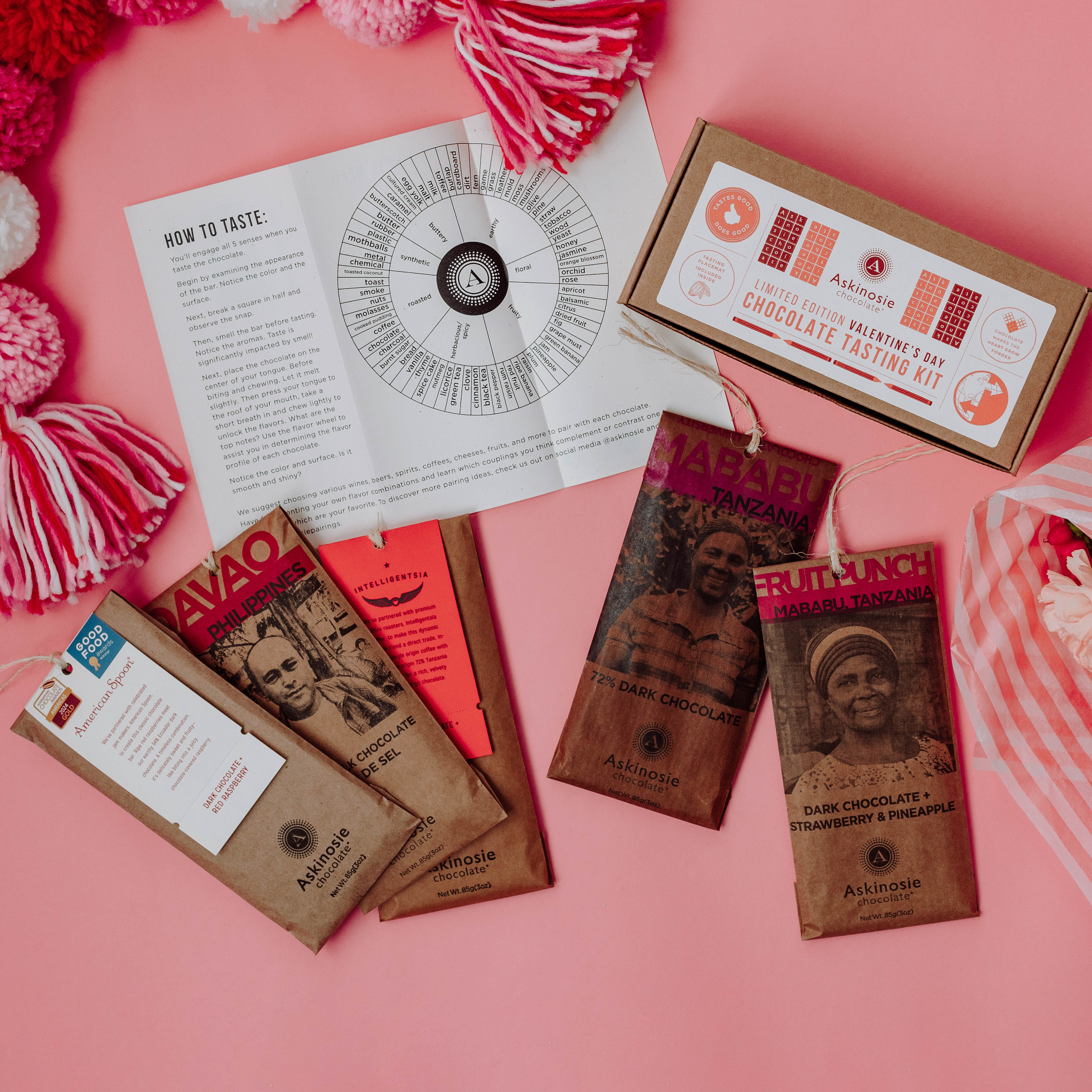 Chocolate Tasting Kit with packaged bars spread out around the kraft paper box and folded tasting placemat on a pink background with flowers nearby.