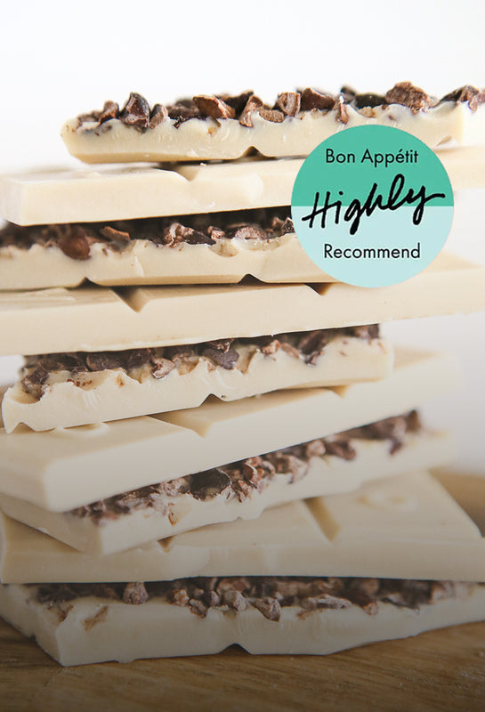 Stacked pieces of a cocoa-nib studded white chocolate bar lay on a wooden board