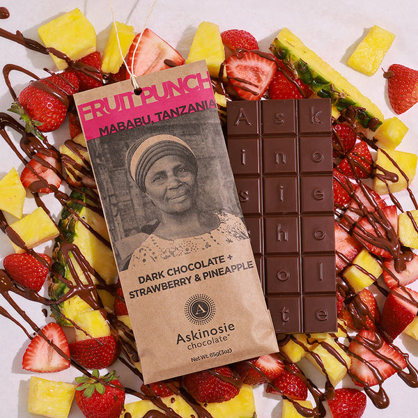 A chocolate bar laid on top of a pile of fresh cut strawberries and pineapples drizzled with melted chocolate