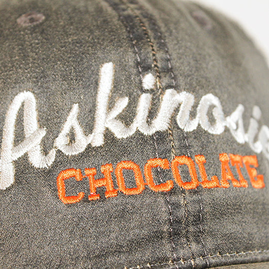 A distressed brown and khaki trucker hat embroidered with Askinosie Chocolate on the front panel